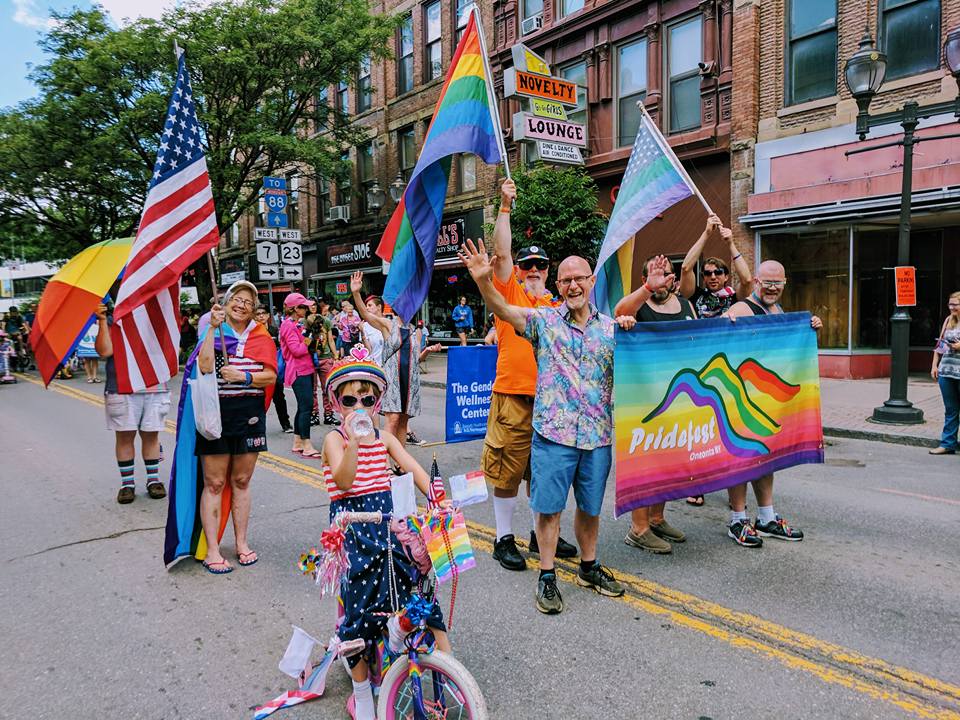 A parade of people of all ages in the street with rainbow signs, flags and clothing celebrating Pridefest in Oneonta, NY